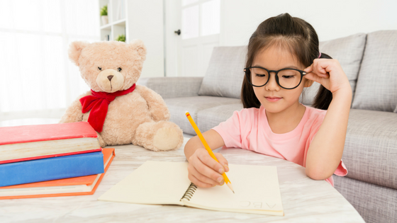 A little girl with glasses is sitting at a table writing in a notebook.